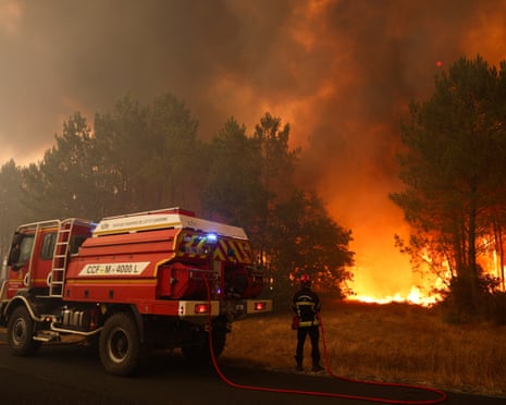 Bright orange fire burns in the background amid some trees as firefighters stand with a hose ready for action