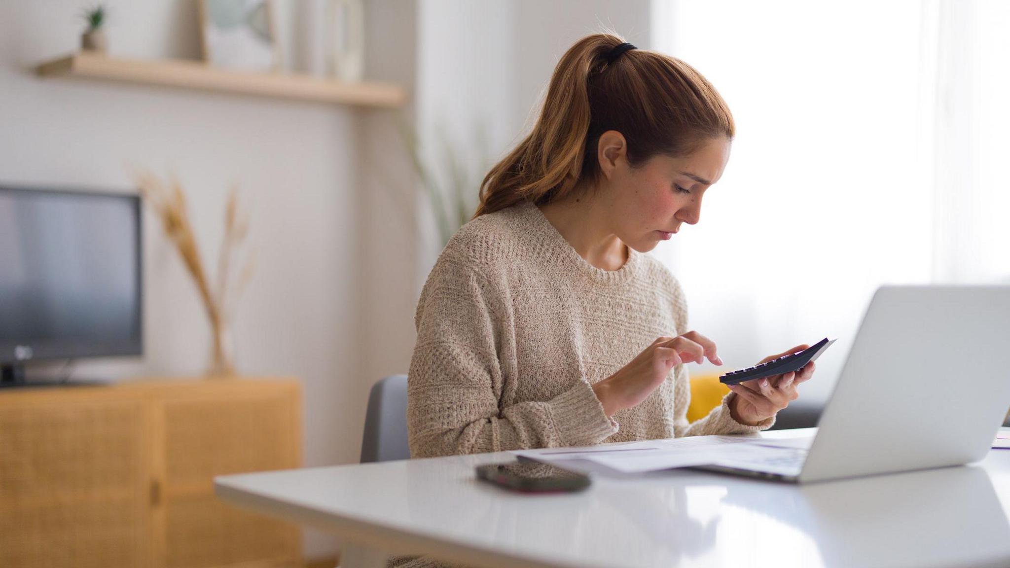 Woman sits at a desk in a home with a phone and a laptop in front of her and a calculator in her hand.